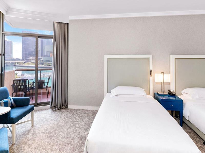 Helles Hotelzimmer mit zwei Einzelbetten, blauem Mobiliar und Balkon mit Stadtblick.