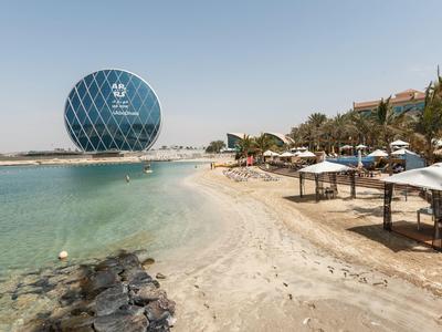 Strand mit Sonnenschirmen und rundem, blauem Hotelgebäude am türkisfarbenen Meer unter klarem Himmel.