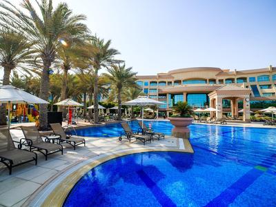 Luxushotel mit großem blauen Pool, Palmen und Liegestühlen bei sonnigem Himmel.
