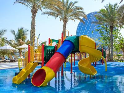 Bunter Wasserspielplatz mit Rutschen im flachen Pool, umgeben von Palmen und Sonnenschirmen.