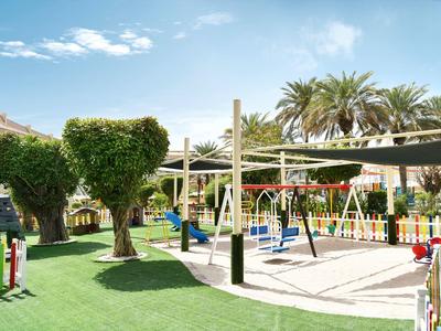 Kinderspielplatz mit Sandfläche, Schaukeln, Rutschen und Bäumen unter blauem Himmel.