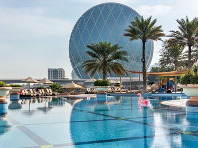 Großer Pool mit Palmen und einem runden modernen Gebäude im Hintergrund bei klarem Himmel.