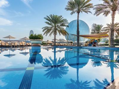 Niebieski basen z palmami i parasolami przed nowoczesną szklaną kopułą pod niebieskim niebem.