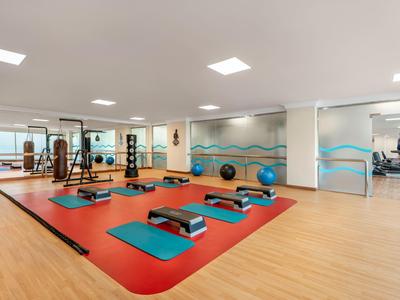 Heller Fitnessraum mit Trainingsmatten, Steppern und Gewichten auf Holz- und rotem Boden.