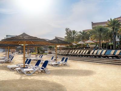 Sandstrand mit Liegestühlen und Sonnenschirmen vor einer Hotelanlage mit Palmen.