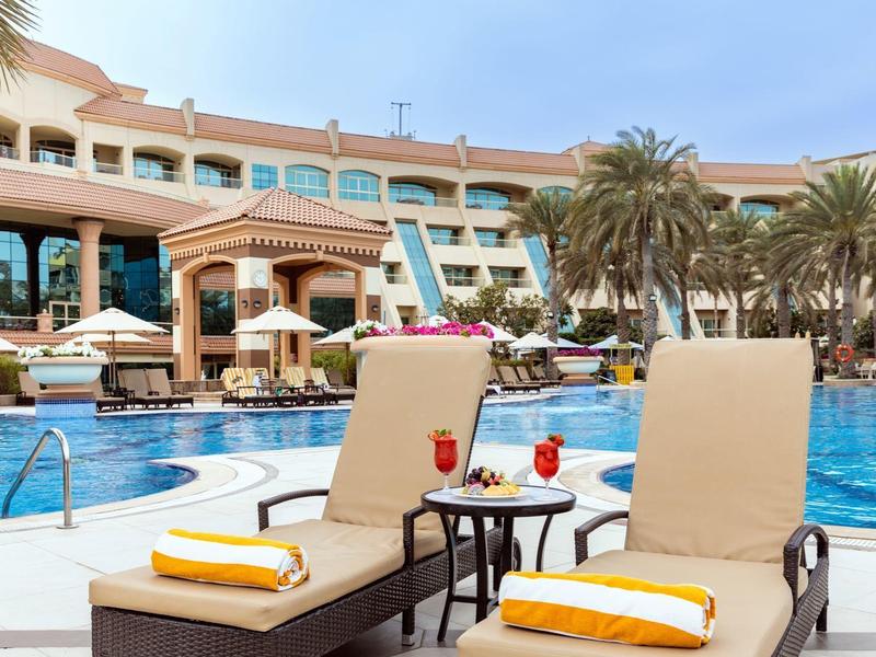 Zwei Poolliegen mit gelben Handtüchern neben einem Pool und Palmen vor einem Hotel.