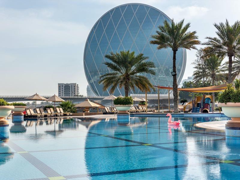 Großer Pool mit Palmen und einem runden modernen Gebäude im Hintergrund bei klarem Himmel.