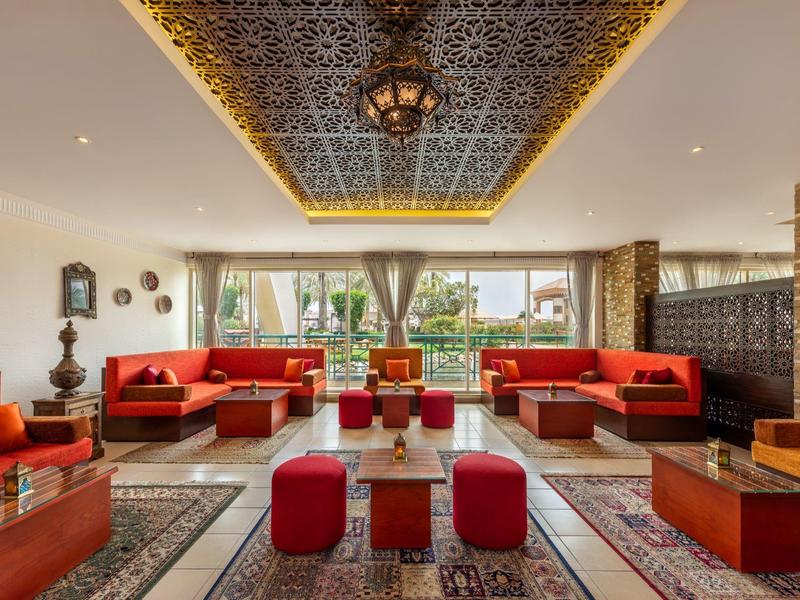 Helles Hotelzimmer mit roten Sitzmöbeln, orientalischen Teppichen und dekorativem Deckendetail.