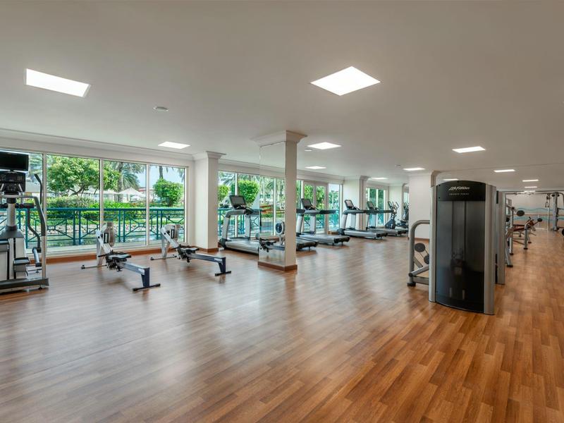 Heller Fitnessraum mit Holzboden, vielseitigen Trainingsgeräten und großen Fenstern mit Gartenblick.