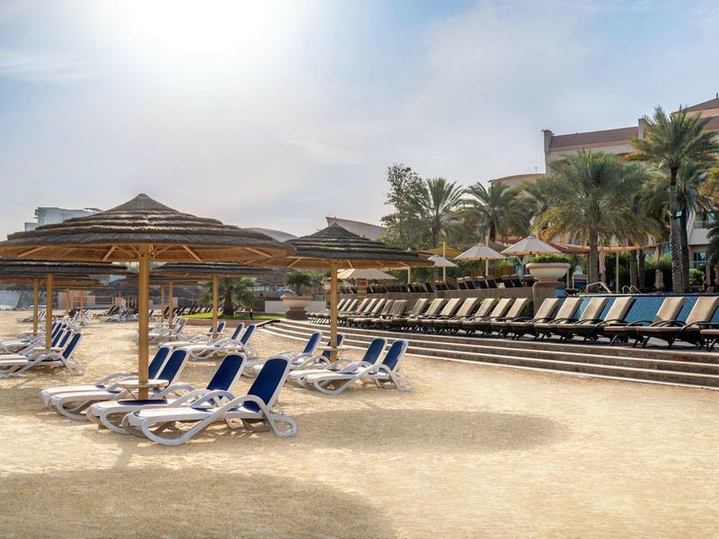 Leere Liegestühle und Sonnenschirme auf einem sonnigen Sandstrand vor einem Hotel mit Palmen.