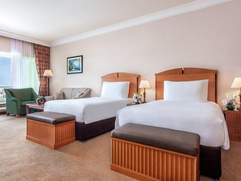 Helles Hotelzimmer mit zwei Doppelbetten, Sesseln und großem Fenster mit Blick nach draußen.