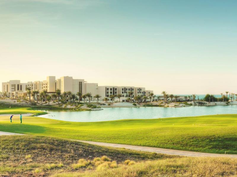 Golfplatz mit grünem Fairway, Teich und Gebäuden im Hintergrund bei klarem Himmel.