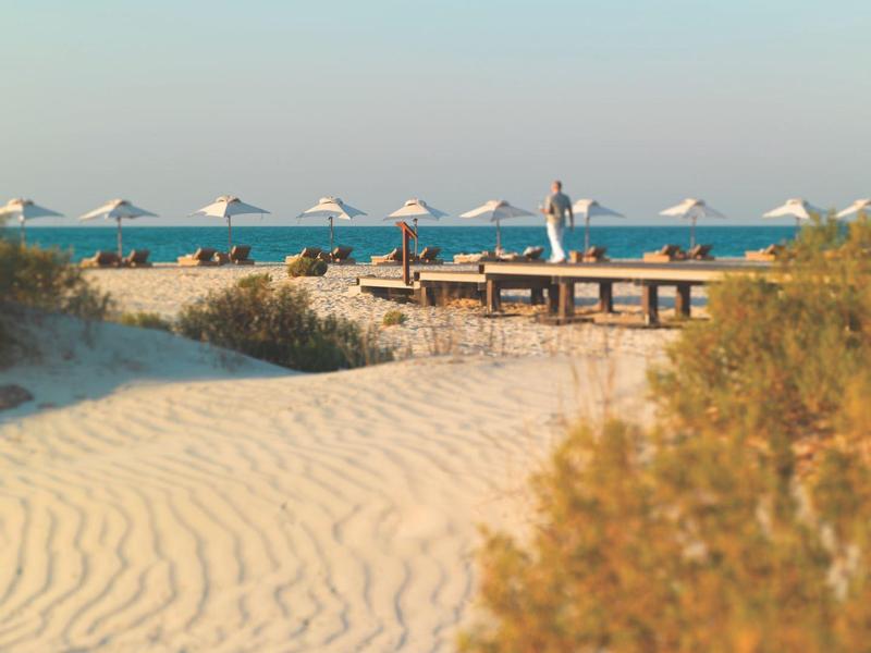 Sandiger Strandweg mit Pflanzen, Liegen und weißen Sonnenschirmen vor ruhigem Meer und klarem Himmel.