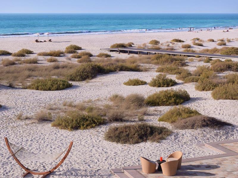 Strand mit Dünen, Sand, blauer Himmel, Hängematte und gemütlicher Sitzgelegenheit mit Kissen.