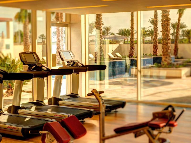 Helles Fitnessstudio mit Laufbändern und Gewichten, große Fenster mit Blick auf Pool und Palmen.