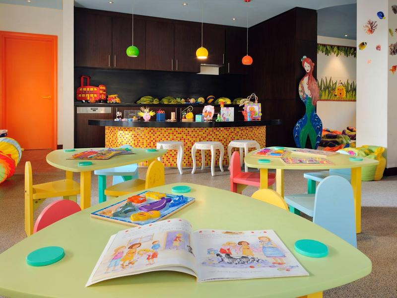 Bunter Kindergartenraum mit Tischen, Stühlen, Bilderbüchern und Regalen voller Spielsachen.