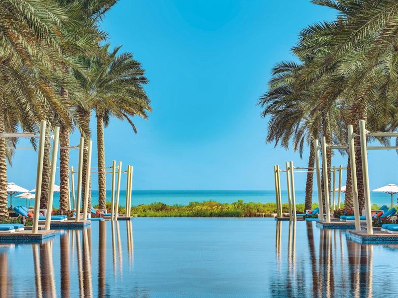 Symmetrischer Pool mit Palmen, blaue Sonne und Meer im Hintergrund, strahlend blauer Himmel.