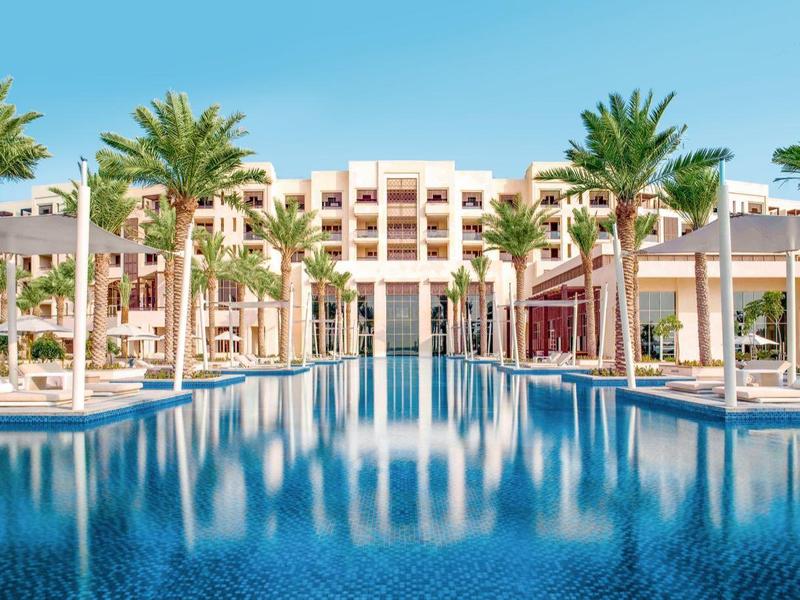 Großer blauer Pool vor modernen, roten Gebäuden mit vielen Palmen und klarem Himmel im Hintergrund.