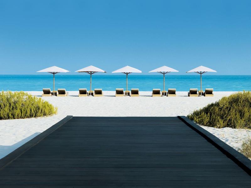 Holzsteg führt zu Strand mit Liegestühlen und weißen Sonnenschirmen vor blauem Meer und Himmel.