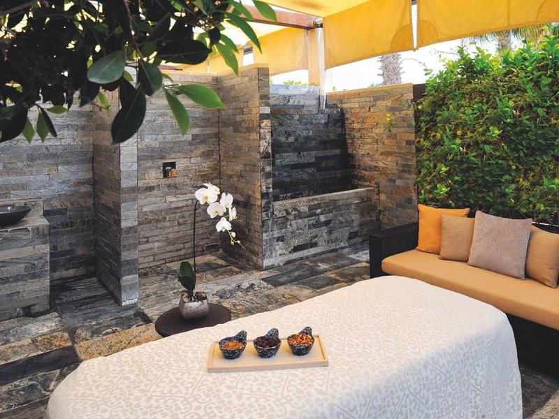 Gemütliche Terrasse mit beiger Couch, weißen Blumen und Wasserfall an grauer Steinwand.