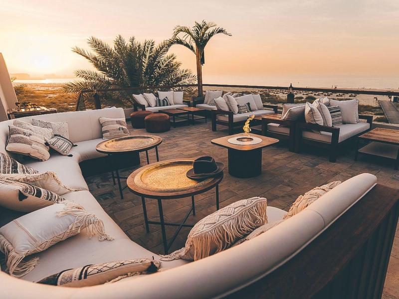 Große runde Lounge mit weißen Polstern, Holztischen und Palmen bei Sonnenuntergang im Freien.