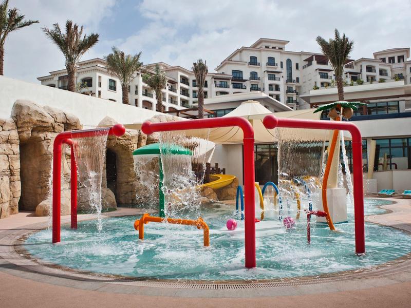 Ein Wasserspielplatz mit roten Gestellen und Wasserfontänen vor einem Hotel mit Palmen.