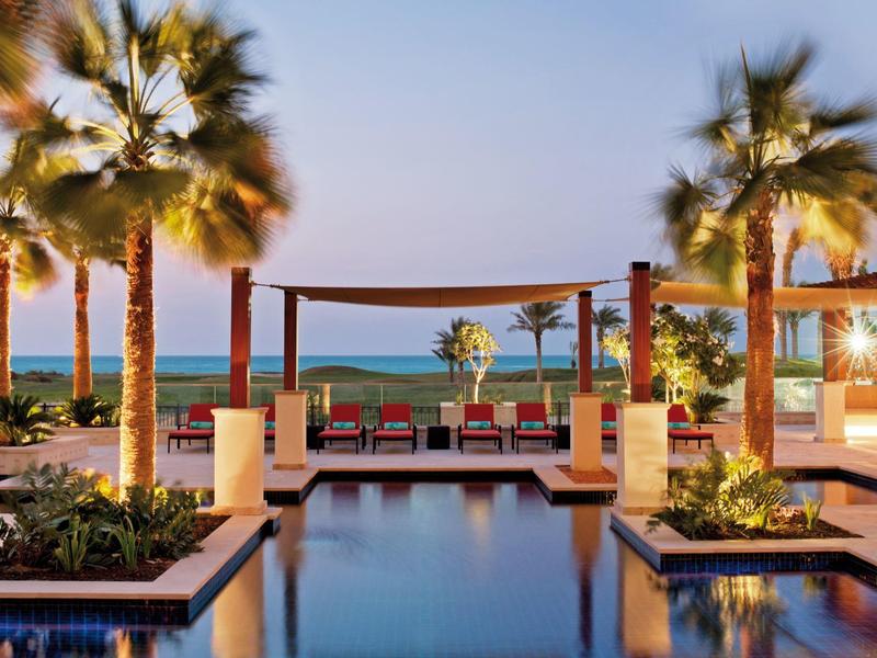 Poolbereich eines Resorts mit Palmen und Blick aufs Meer bei Sonnenuntergang.