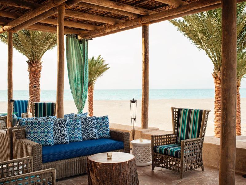 Überdachte Lounge mit blauen Kissen, Holzmöbeln und Palmen mit Blick auf den Strand.
