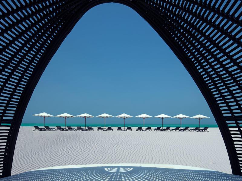 Strandliegen mit weißen Sonnenschirmen am klaren blauen Meer, gesehen durch eine schwarze Struktur.