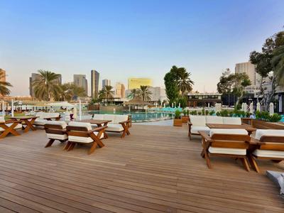 Große Holzterrasse mit bequemen Sitzmöbeln neben einem Pool in einer urbanen Hotelanlage.