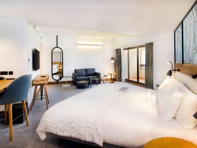 Helles Hotelzimmer mit großem Bett, Schreibtisch, Sessel und Balkon mit Blick nach draußen.