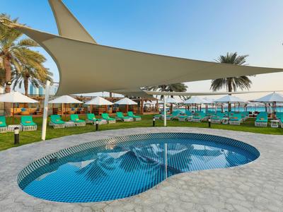 Poolbereich mit Ligstühlen und Sonnensegeln neben dem Meer in einem Resort.