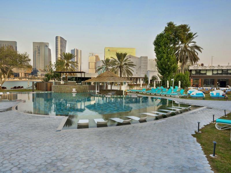 Moderner Außenpool mit Liegestühlen und Palmen vor einer Stadt-Skyline bei klarem Himmel