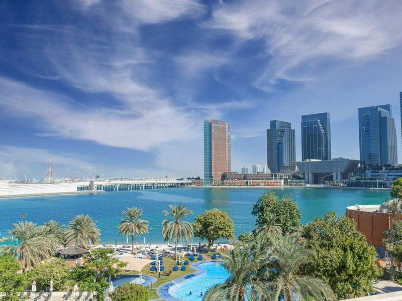 Blick auf eine Hotelanlage mit Pool, Palmen und einem modernen Stadtbild am Wasser.