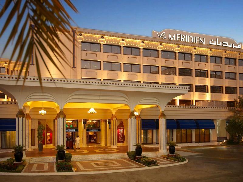 Hotel mit großem Eingangsbereich, gelber Beleuchtung und Palmen vor dem Gebäude.