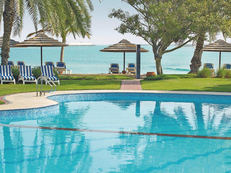 Blick auf einen Pool mit Liegestühlen und Sonnenschirmen am Meer in einem Hotelgarten.