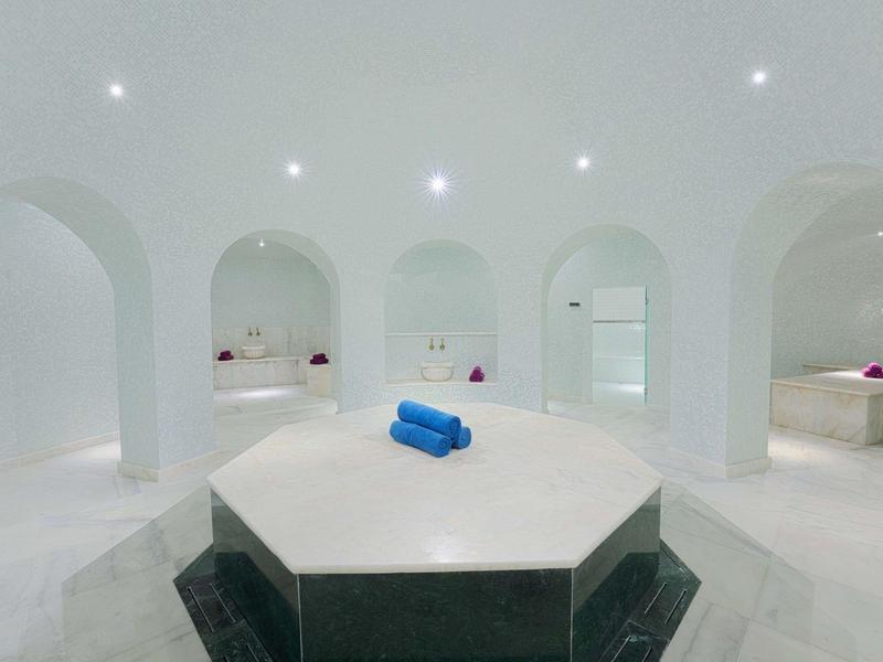 Heller, weißer Spa-Bereich mit runden Bögen und zentralem Marmorruhetisch mit blauen Handtüchern.