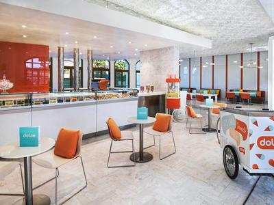 Helle Hotel-Lounge mit weißen Tischen, orangen Stühlen und einem Eiswagen.