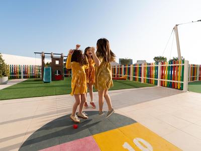 Drei Mädchen in gelben Kleidern spielen auf einem sonnigen, farbenfrohen Spielplatz eines Hotels.
