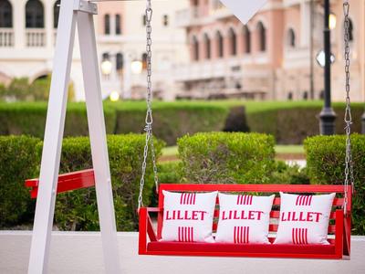Rote Schaukel mit Lillet-Logo vor einem historischen Gebäude und grünen Sträuchern.