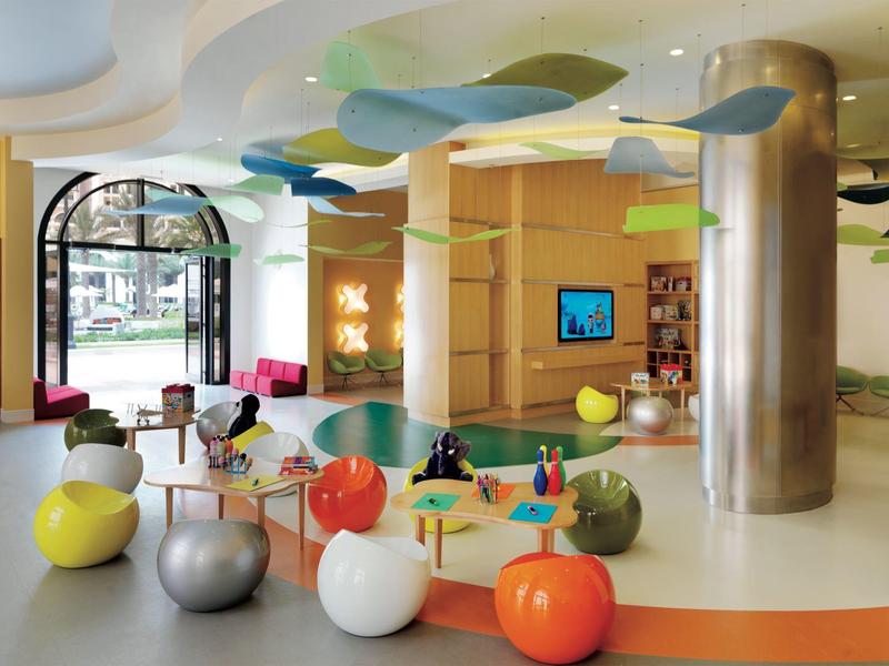 Area giochi colorata per bambini con palloni, tavoli e televisore in una stanza moderna.