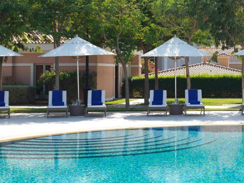 Piscina con lettini blu e ombrelloni bianchi in un giardino d'hotel.