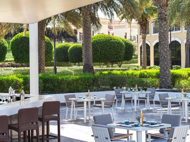 Ristorante all'aperto con tavoli, sedie e vista su un giardino curato con palme.