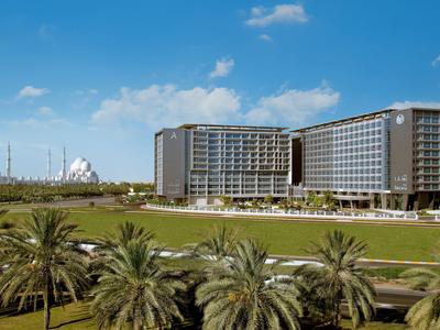 Modernes Hotel mit zwei großen Gebäuden, umgeben von Palmen und grüner Wiese unter blauem Himmel.