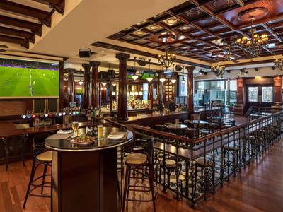 Elegant hotel bar with wooden floors, bar stools, large windows, and a wall-mounted TV.