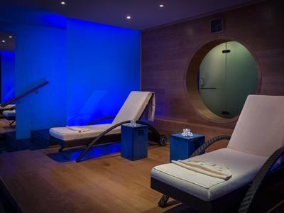 Relaxation room with white loungers, blue-lit background, and a round window in the wall.