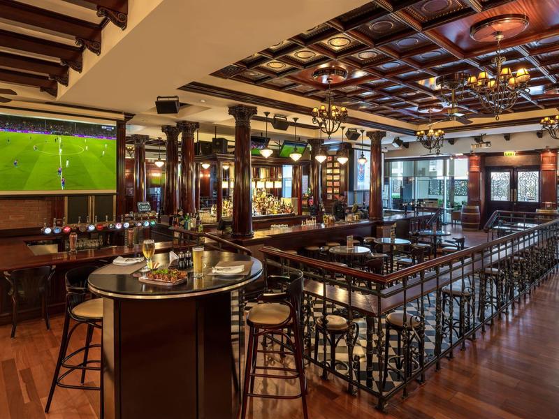 Elegant hotel bar with wooden floors, bar stools, large windows, and a wall-mounted TV.