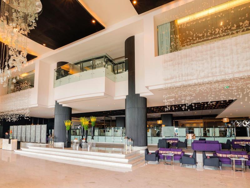 Moderní hotelová lobby s recepcí, posezením, velkými lustry a otevřenou galerií.