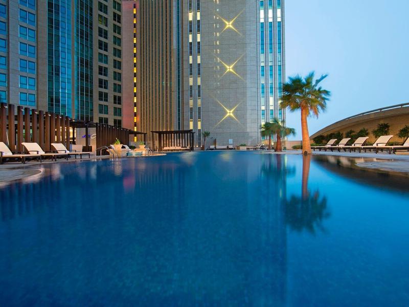 Moderní venkovní bazén s lehátky a palmami vedle vysokých budov za soumraku.