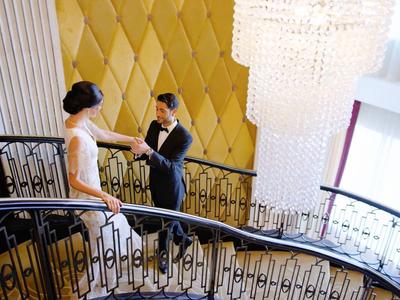 Ein Paar steigt eine elegante, geschwungene Treppe mit schwarzem Geländer und goldenem Wandmuster hinauf.